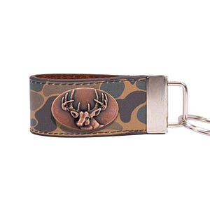 May include: A camouflage pattern leather keychain with a copper-colored oval emblem featuring a deer head. The keychain has a silver-colored metal clasp and a key ring. The leather is brown on the inside.