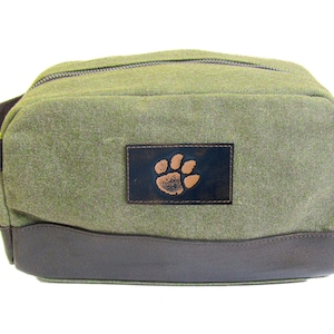 Könnte beinhalten: Eine grüne Reisetasche aus Canvas mit braunem Lederbesatz und einem braunen Lederflicken mit einem Pfotenabdruck.