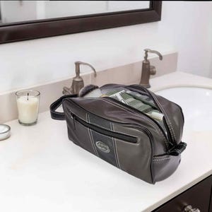 Puede incluir: Un neceser de cuero gris oscuro con asa y cremallera negras. La bolsa est&aacute; abierta, revelando un vistazo de su contenido. La bolsa est&aacute; sobre una encimera blanca en un ba&ntilde;o.