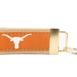 May include: A tan and orange key chain with a white Longhorn steer logo. The key chain has a gold metal ring and a gold metal clasp.