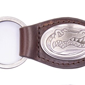 May include: Brown leather keychain with a silver oval emblem featuring a detailed alligator design. The keychain includes a silver key ring. The leather is stitched around the emblem.