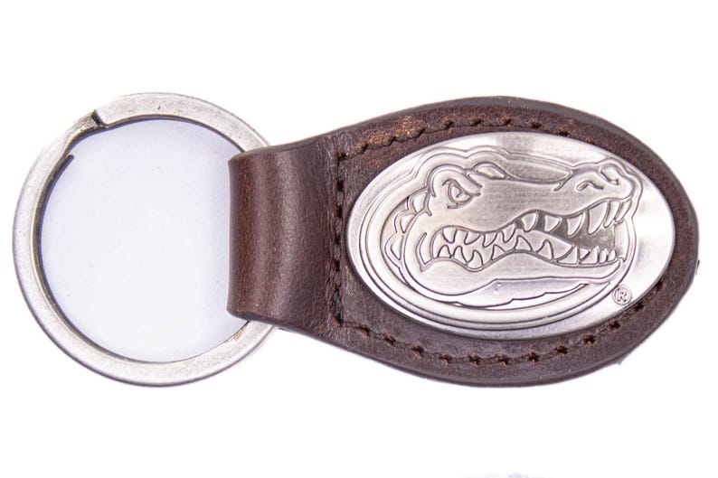 May include: Brown leather keychain with a silver oval emblem featuring a detailed alligator design. The keychain includes a silver key ring. The leather is stitched around the emblem.