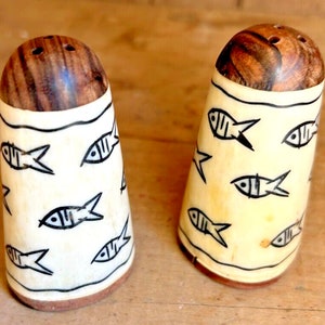 Puede incluir: Un par de saleros y pimenteros. Los saleros tienen una base beige claro con ilustraciones de peces negros. Las tapas son de madera marrón oscuro. Los saleros son cilíndricos y tienen un aspecto rústico y hecho a mano.