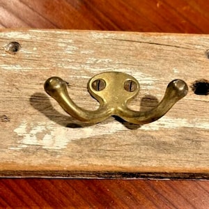 May include: A rustic wooden wall hook with a double-pronged brass hook. The wood is weathered and has a white painted design.