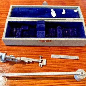 May include: A silver drafting compass with a blue velvet lined case. The compass has a knob on the end of the arm and a small dial with the number 17013 on it. The case has a silver latch.