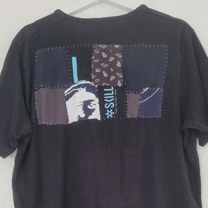 Op de afbeelding: Zwart T-shirt met een patchwork-ontwerp op de rug. Het patchwork omvat verschillende stoffen en kleuren, waaronder blauw, grijs en zwart. De tekst "#SKILL" is zichtbaar op het patchwork.