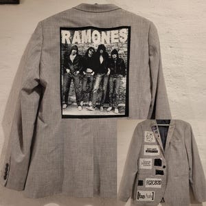 Op de afbeelding: Een grijze blazer met een Ramones-afbeelding op de rug. De blazer heeft een klassiek ontwerp met een gekerfde revers en knoopsluiting. Er zijn patches op de voorkant genaaid. De blazer is maat medium.