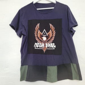 Può includere: Una maglietta blu navy con una grafica in bianco e nero di un'aquila con il testo "MEGA BABE" e "MIDNIGHT RIDE". La maglietta ha un fondo a righe verde e nero.