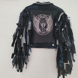 Puede incluir: Chaqueta vaquera oscura con mangas de tela deshilachada. La espalda presenta un parche negro con un emblema de serpiente alada y las palabras "PURE METAL" y "STAY BOLD". El cuello está tachonado.
