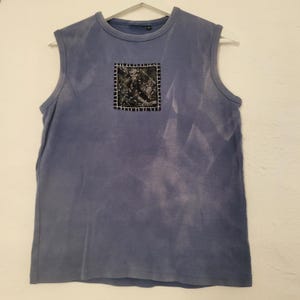 May include: A sleeveless, blue-grey top with a square patch on the front. The patch is black with white stitching and a textured design. The top is hanging on a white hanger.