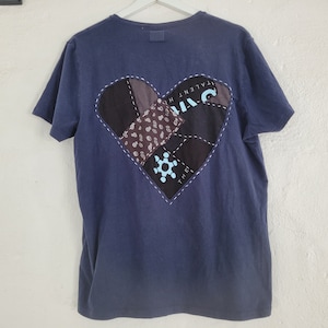 Può includere: Una t-shirt blu navy con una toppa a forma di cuore cucita sul retro. La toppa è realizzata con diversi tessuti e presenta il testo "TALENT HT" e "THE".