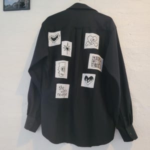 May include: Black button-down shirt with several white patches, each displaying a different black illustration. The patches include a bat, a star, a heart, a cartoon character, and the text "she wants revenge".