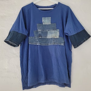 Op de afbeelding: Een blauw T-shirt met een groot denim patch-ontwerp op de voorkant. De patch is gemaakt van verschillende stukken denim die in een gelaagd patroon aan elkaar zijn genaaid.