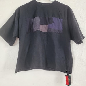 Op de afbeelding: Zwart T-shirt met korte mouwen met een patchwork ontwerp in tinten paars, grijs en blauw over de borst. Een klein, decoratief item hangt aan de onderste zoom. Het shirt heeft een rauwe, onafgewerkte zoom.
