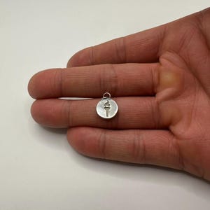 Op de afbeelding: Een kleine, ronde zilveren hanger met een fakkelontwerp. De hanger wordt in een hand gehouden tegen een witte achtergrond. De fakkel is afgebeeld met een vlam aan de bovenkant.