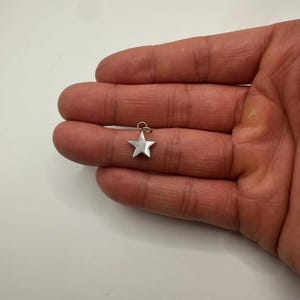 Puede incluir: Un pequeño dije de estrella plateada con un lazo en la parte superior. La estrella de cinco puntas se muestra sobre un fondo neutro. Este dije es adecuado para la fabricación de joyas o como colgante.