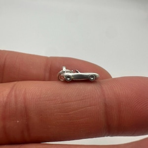 May include: A small, silver-colored metal charm shaped like a convertible sports car. The charm features a smooth, polished finish with defined wheel wells and a small loop at the front for attachment to a bracelet or necklace chain.