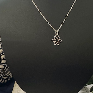 Pode incluir: Um colar prateado com um pendente em forma de átomo. O pendente apresenta um design delicado e vazado. O colar é exibido em um manequim preto.