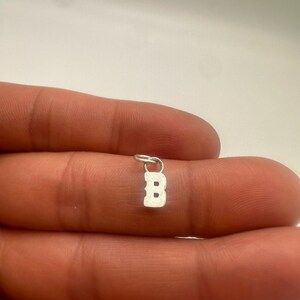 Puede incluir: Un pequeño dije de metal de color plateado con la forma de la letra mayúscula B. El dije presenta un estilo de fuente clásica con remates e incluye una pequeña argolla circular unida en la parte superior para colgarlo en una cadena.