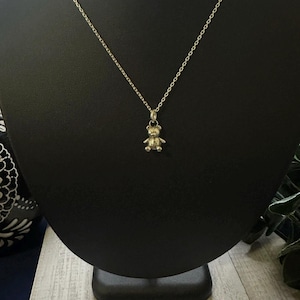 May include: A gold-colored necklace featuring a small teddy bear pendant. The pendant is detailed with visible arms, legs, and a head. The necklace is displayed on a black jewelry stand.