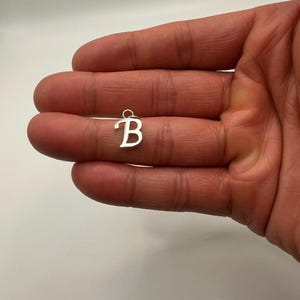 Pu&ograve; includere: Un piccolo ciondolo color argento a forma di lettera "B" in corsivo. Il ciondolo ha un piccolo anello nella parte superiore per il fissaggio a una catena o un braccialetto. Lo sfondo &egrave; di colore chiaro e neutro.