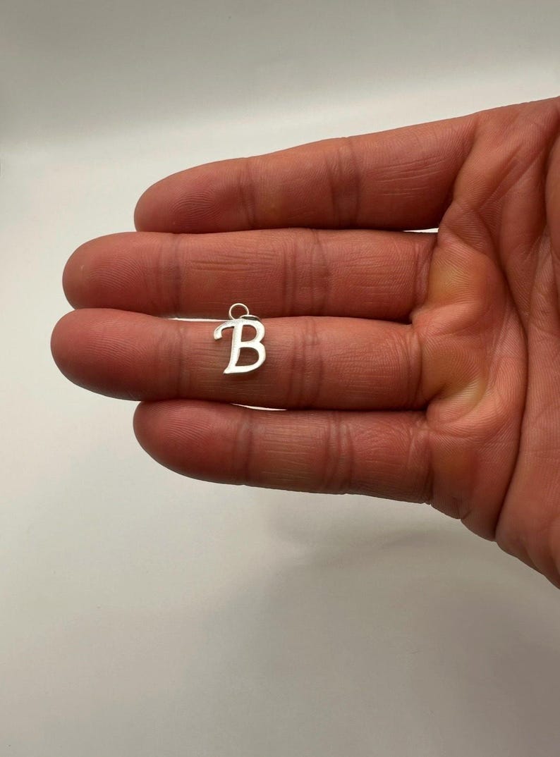 Pu&ograve; includere: Un piccolo ciondolo color argento a forma di lettera "B" in corsivo. Il ciondolo ha un piccolo anello nella parte superiore per il fissaggio a una catena o un braccialetto. Lo sfondo &egrave; di colore chiaro e neutro.