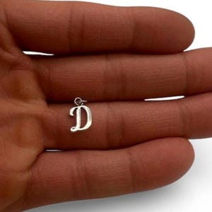 May include: A small, silver-colored metal charm in the shape of the uppercase letter D. The letter features a classic, serif-style font with a small circular jump ring attached to the top, allowing it to be suspended from a chain or bracelet.