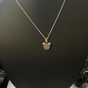 May include: Silver necklace with a miniature clock pendant. The pendant has a detailed clock face and two tiny bells. The necklace is displayed on a black mannequin, highlighting the jewellery's design and style.