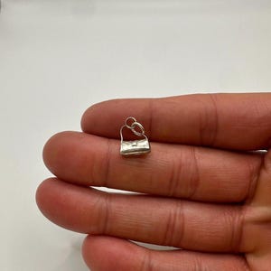 May include: A small, silver-coloured metal charm shaped like a miniature handbag. The charm features a rectangular body with a structured flap and a thin, arched handle attached to a circular jump ring at the top for hanging on a chain or bracelet.