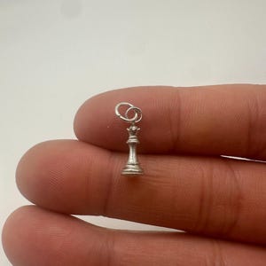 May include: A small silver-colored metal charm shaped like a chess queen piece. The charm features a detailed crown top and a flared base, with a small circular jump ring attached to the top for hanging. The piece has a matte, metallic finish.