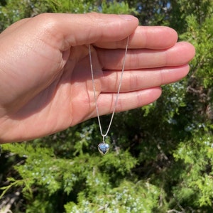 May include: A silver chain necklace with a small, heart-shaped pendant. The necklace is held in a hand against a blurred background of green foliage.