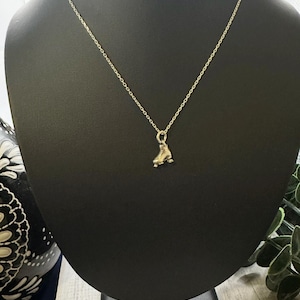 Pode incluir: Um colar dourado com um pequeno pingente de patins. O pingente tem rodas e cadarços detalhados. O colar é exibido em um busto preto sobre um fundo neutro.
