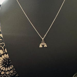 May include: A silver-colored necklace with a small rainbow pendant. The necklace is displayed on a black mannequin. The rainbow pendant is a simple, arched design.