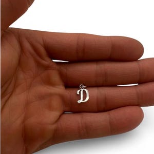 May include: A small, silver-colored pendant in the shape of the letter "D". The pendant is displayed against a neutral background. The letter has a simple, elegant design, with a small loop at the top for attaching to a chain or bracelet. The pendant is likely made of sterling silver.