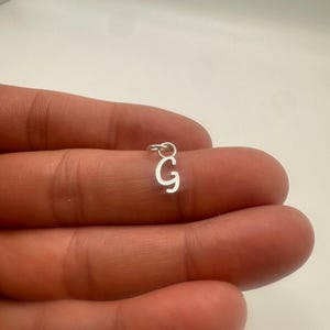Könnte beinhalten: Ein kleiner, silberfarbener Metallanhänger in Form des Großbuchstabens G. Der Anhänger hat eine schlichte, klare Schriftart und verfügt über einen kleinen, runden Biegering an der Oberseite zum Aufhängen oder Befestigen an einer Kette.