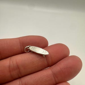 May include: A small silver-colored skateboard charm featuring a smooth, elongated deck with four tiny drilled holes on each end and a jump ring attached to one side for hanging. The miniature skateboard has visible wheels underneath.