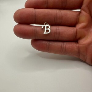 Pu&ograve; includere: Un piccolo ciondolo color argento a forma di lettera "B". Il ciondolo &egrave; tenuto in una mano su uno sfondo bianco. La lettera &egrave; in un semplice carattere sans-serif e ha un piccolo anello nella parte superiore.