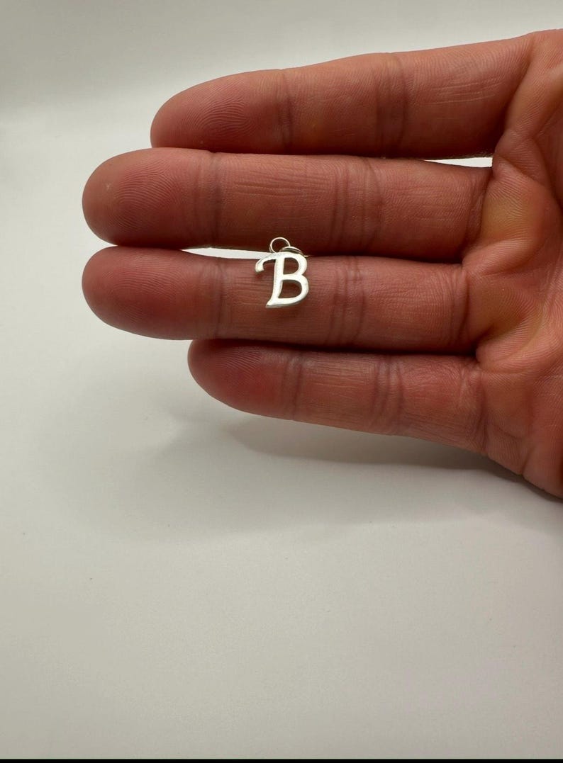 Pu&ograve; includere: Un piccolo ciondolo color argento a forma di lettera "B". Il ciondolo &egrave; tenuto in una mano su uno sfondo bianco. La lettera &egrave; in un semplice carattere sans-serif e ha un piccolo anello nella parte superiore.