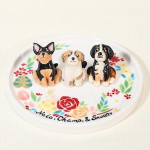 Puede incluir: Un plato de cerámica blanco con un diseño floral y tres figuras de perros. Los perros son negros, marrones y blancos. El plato tiene el texto "Nala, Champ, & Snoopy" en una fuente manuscrita.