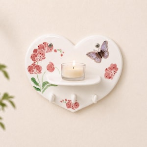 May include: A white heart-shaped ceramic wall shelf featuring a painted design of pink orchids and a purple butterfly. The shelf includes a small glass votive candle holder with a lit white candle and three white hooks at the bottom for hanging.