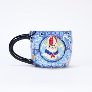May include: A ceramic mug with a black handle and a blue exterior. The mug features a whimsical design of a seal wearing a blue scarf and a red and white striped ball on its head. The background is decorated with blue waves and stars.
