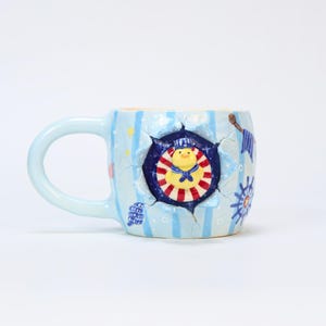 May include: A light blue ceramic mug with a handle, decorated with vertical stripes, stars, and nautical-themed illustrations. The mug features a central design of a sailor duck in a circular frame, with red and white stripes. The interior is a light yellow colour.
