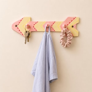 May include: A decorative wooden arrow-shaped wall hook in pink and yellow. The arrow has three small hooks, holding keys, a light blue shirt, and a pink scrunchie. The design features a dashed red stitch pattern.