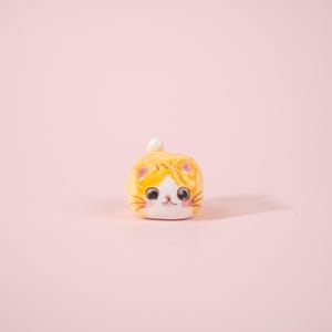 Peut inclure: Petite figurine de chat en céramique jaune et blanche, au design mignon et cartoon. Le chat présente des marques oranges, un visage blanc et une petite boule blanche sur la tête. La figurine est posée sur un fond rose tendre.