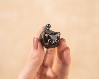 Könnte beinhalten: Kleine, glänzend schwarze Katzenfigur mit rosa Akzenten an Ohren und Wangen. Die Katze hat weiße Schnurrhaare und einen kleinen, aufgestellten Schwanz. Die Figur wird in einer Hand vor neutralem Hintergrund gehalten.