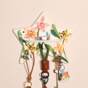 May include: A star-shaped key holder with a floral design in orange, yellow, and pink. It has a small shelf with a lit candle and three hooks holding keychains. The keychains have leather and fabric accents.
