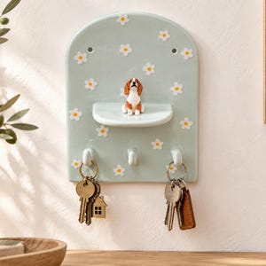 May include: A light green ceramic wall-mounted key holder with an arched top, decorated with white daisy patterns. It features a small central shelf topped with a miniature beagle figurine and three hooks at the bottom holding metal keys.