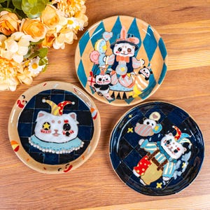 Può includere: Tre piatti in ceramica con disegni dipinti a mano che raffigurano panda vestiti da clown. I piatti hanno un motivo a scacchi blu e neri con un bordo beige.