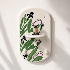 Può includere: Un organizer da parete ovale in ceramica con un design dipinto a mano di foglie verdi, fiori di iris blu scuro e mughetti viola. Include una piccola mensola centrale con una figura di gatto scolpita e tre ganci bianchi.