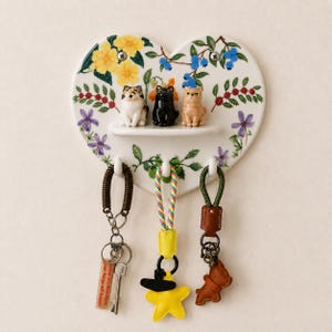 May include: A heart-shaped wall-mounted key holder with a floral pattern featuring yellow flowers and blue berries. It includes a small shelf holding three miniature cat figurines and three white hooks below, each supporting a keychain with various charms.